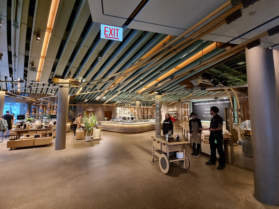 Interior of a modern coffee shop with a high ceiling. Three staff members stand near a cart. An "EXIT" sign hangs above.