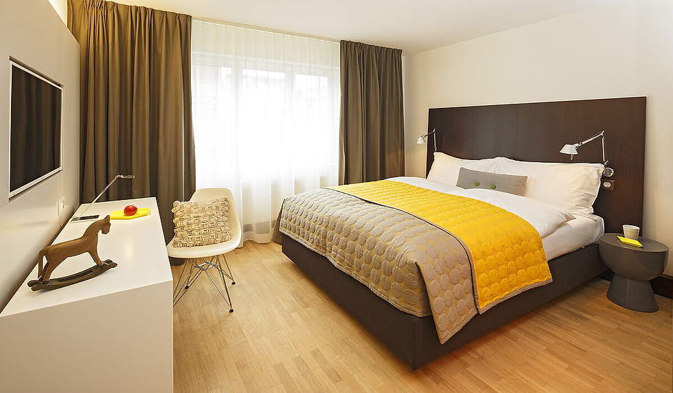 Modern bedroom at Hotel Rossli Zurich, with a bed, yellow quilt, desk, chair, TV, and window with curtains; bright, tidy, minimalist mood