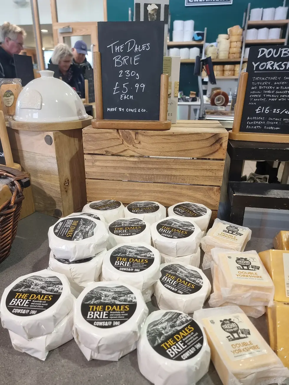 Brie cheese display at wensleydale creamery with labelled "The Dales Brie £5.99" sign on a wooden counter. Customers browse in the background, cheeses stacked neatly.