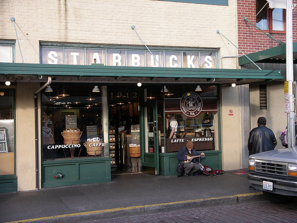 Man plays guitar outside the original Starbucks with a green facade. Another person stands nearby. The store displays coffee signs in the window.