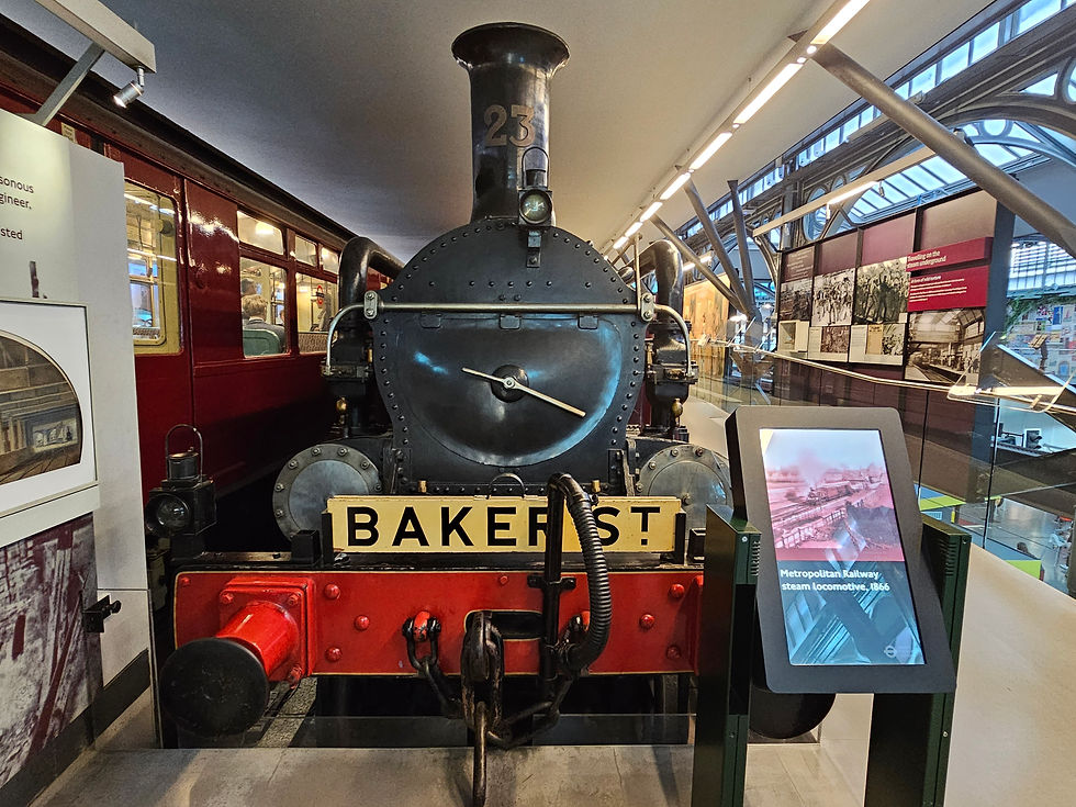 Historic steam locomotive with "BAKER ST" sign, red and black colors, displayed indoors at the London Transport Museum. Nearby, an info screen reads "Metropolitan Railway, 1866."