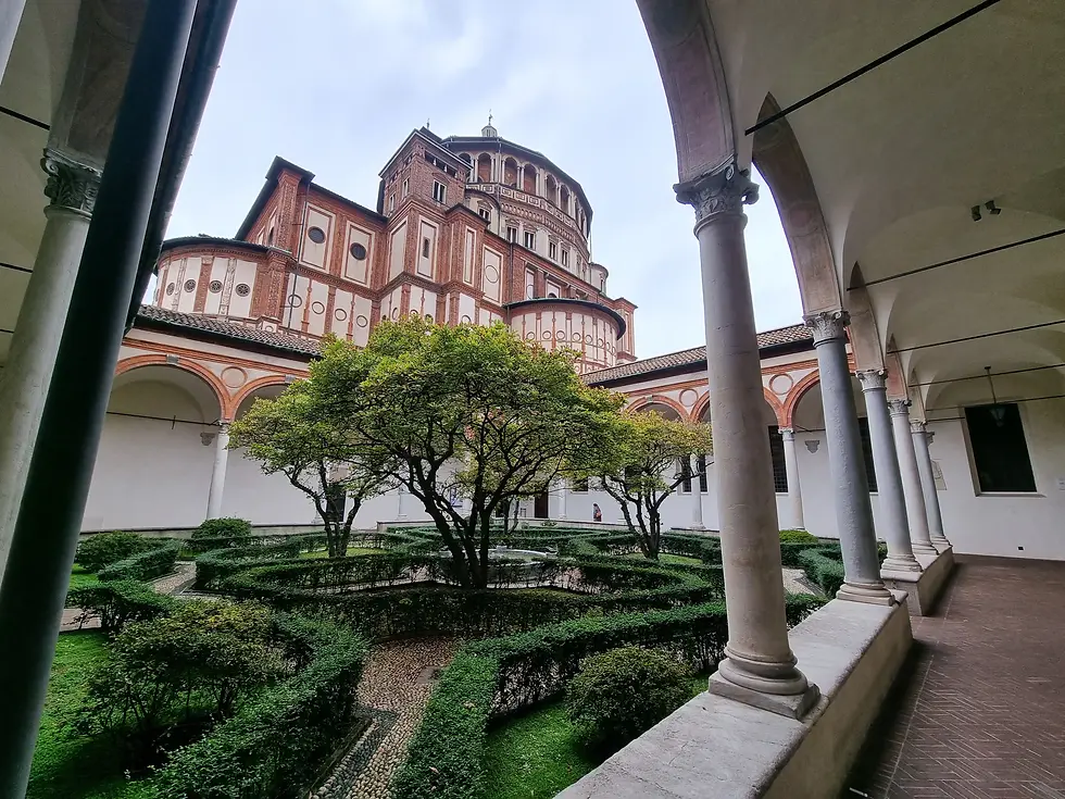 Cloister garden with hedges and a central tree, surrounded by arches and a historic brick building. Overcast sky sets a calm mood.
