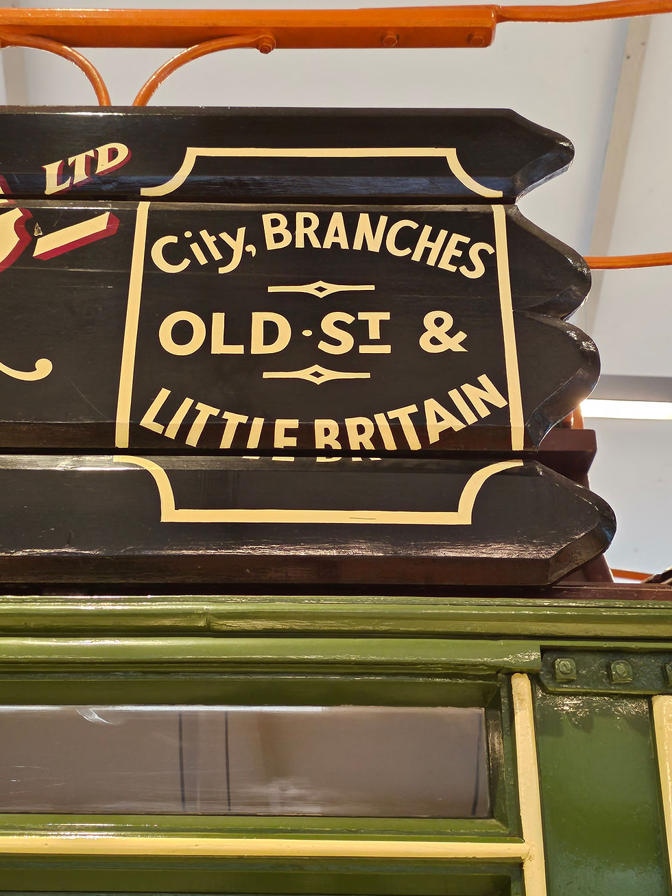 Close-up of a vintage black and cream sign with text: "City, Branches, Old St & Little Britain," above a green frame. This signwriting is on an exhibit bus at the London transport Museum