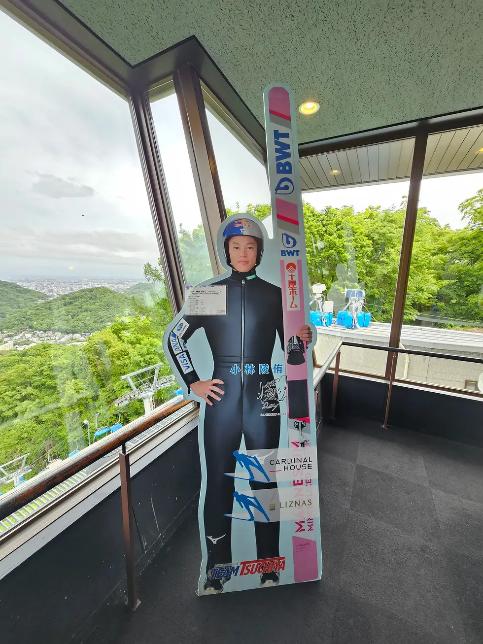 Cardboard cutout of a ski jumper with pink and blue skis. Set in a glass-paneled room with a view of lush green hills and cloudy sky over the Ōkurayama Ski Jump in Sapporo.