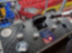Car simulation control panel with grip handles, buttons, and text about practicing wheel changes at the Silverstone museum. Red and blue vehicle parts in the background.