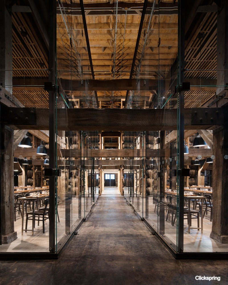 Industrial interior with wooden beams, glass walls, and dining tables. Dimly lit with a warm, rustic feel. "Clickspring" text in corner.
