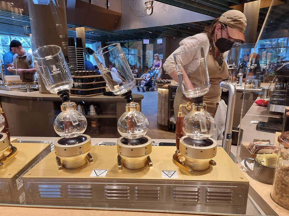 Coffee shop scene with a masked barista using siphon coffee makers on a counter. Warm lighting and a busy background with patrons.
