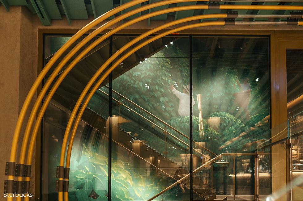 Modern interior with green foliage mural behind glass staircase. Yellow pipes curve overhead. "Starbucks" text in corner. Warm lighting.