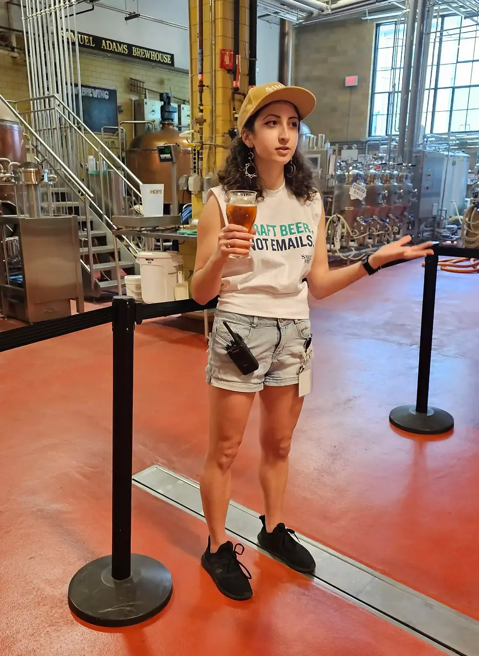 A person in casual attire holds a beer inside the brewhouse at Samuel Adams Boston Brewery Tour. Background shows brewing tanks. Shirt text: "Draft Beer, Not Emails."