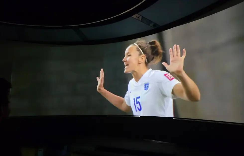 Women’s soccer player in white jersey number 15, raising both hands and smiling on a large screen, with England crest visible at the FIFA Museum.