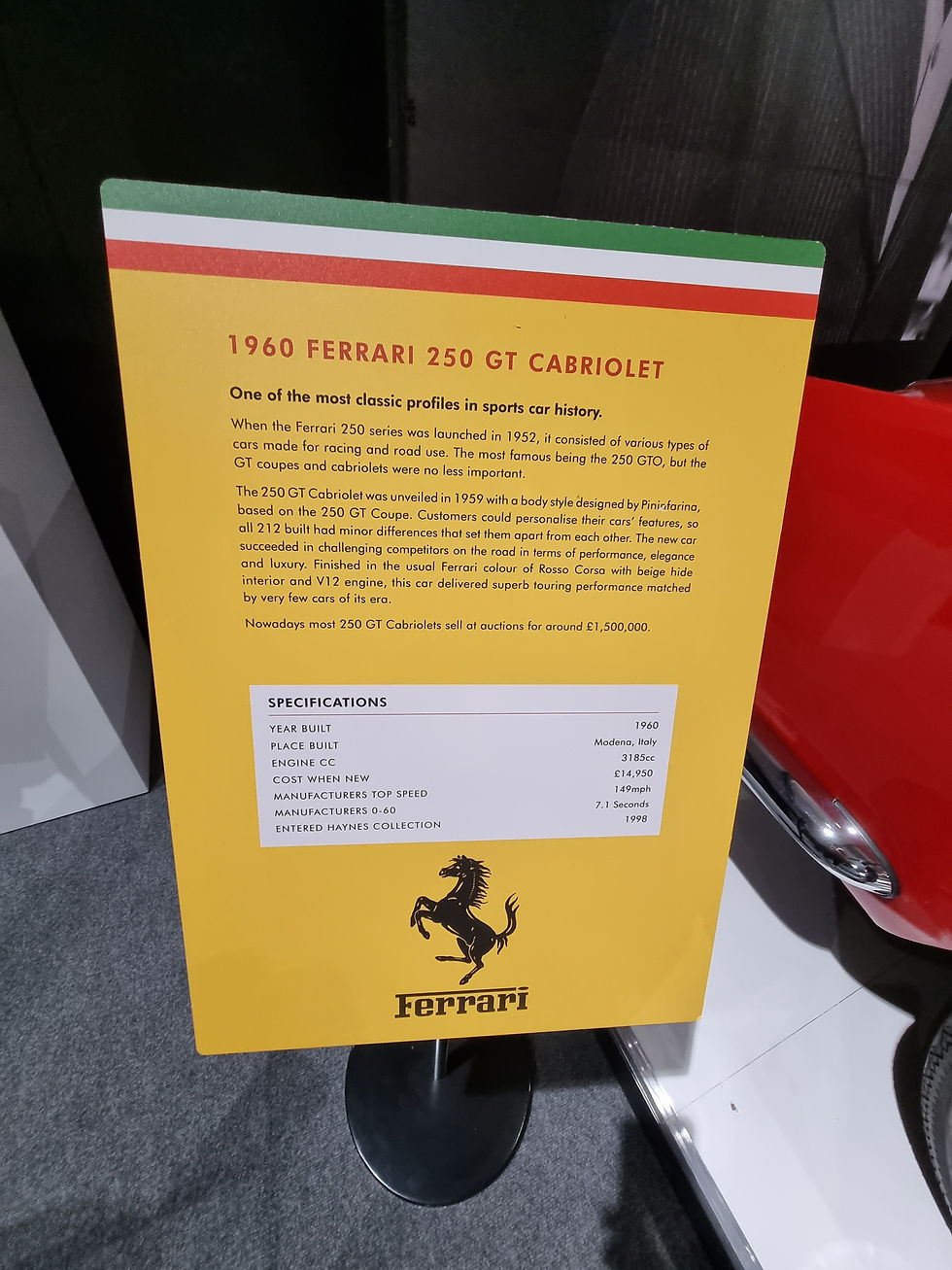 Sign at the Haynes Motor Museum detailing the 1960 Ferrari 250 GT Cabriolet, yellow background, specs like engine, top speed, and cost. Red car visible beside it.