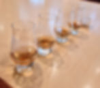 Four whiskey glasses on a bar counter with amber liquid. Background has muted lighting and TV screens. Cozy atmosphere.