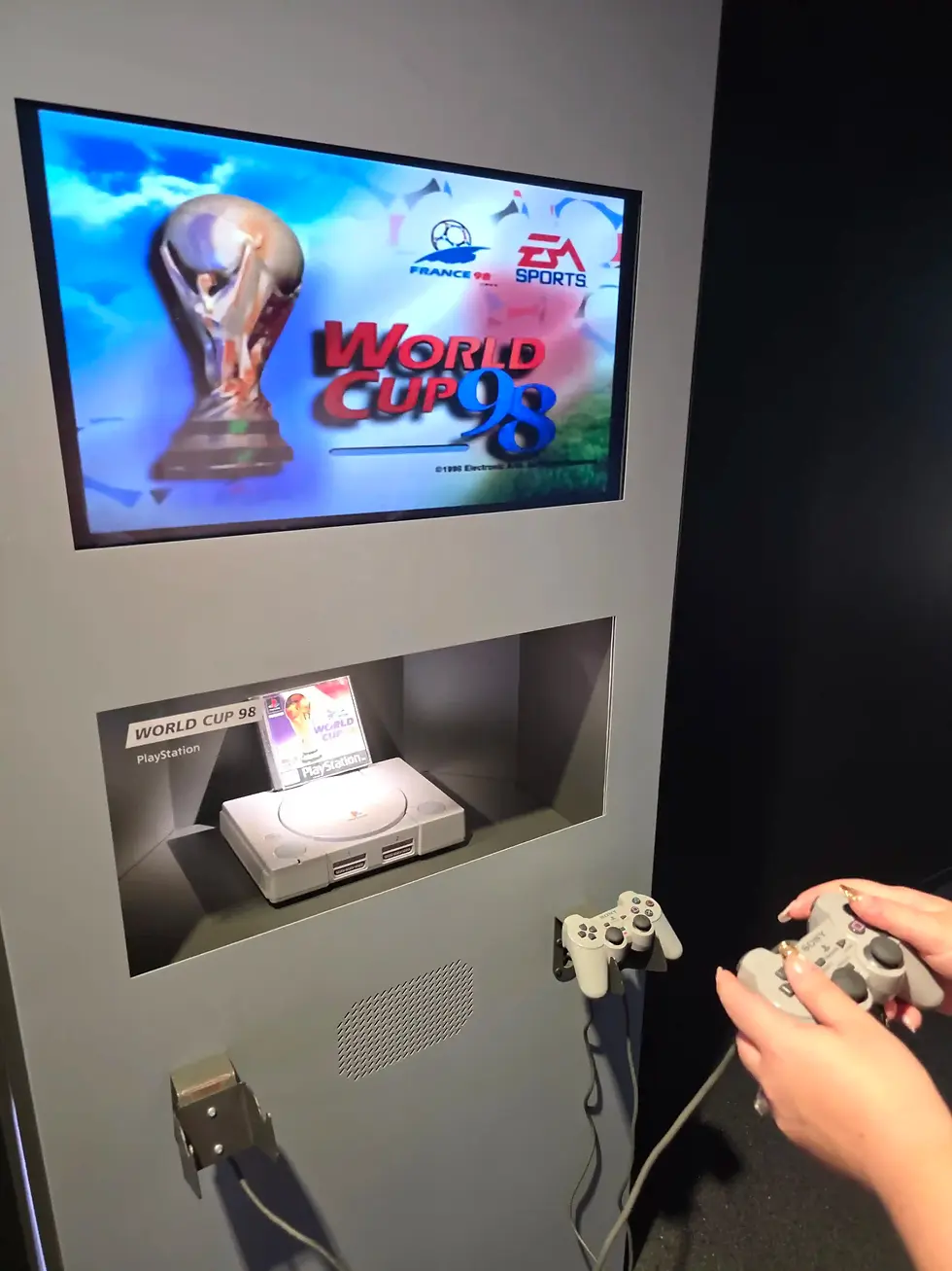 Display showing World Cup 98 on a PlayStation demo, with hands holding gray controllers beside the console display at the FIFA Museum.