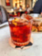 A vibrant red negroni cocktail with ice and orange peel sits on a coaster on a marble table. Blurred cafe setting in the background.