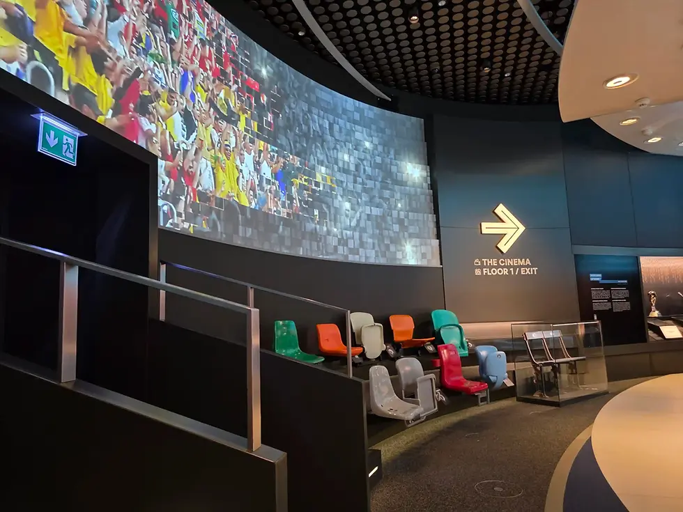 Modern cinema exhibit at FIFA museum with colorful stadium seats, large cheering crowd screen, and exit sign pointing to Floor 1 in a dark museum-like space