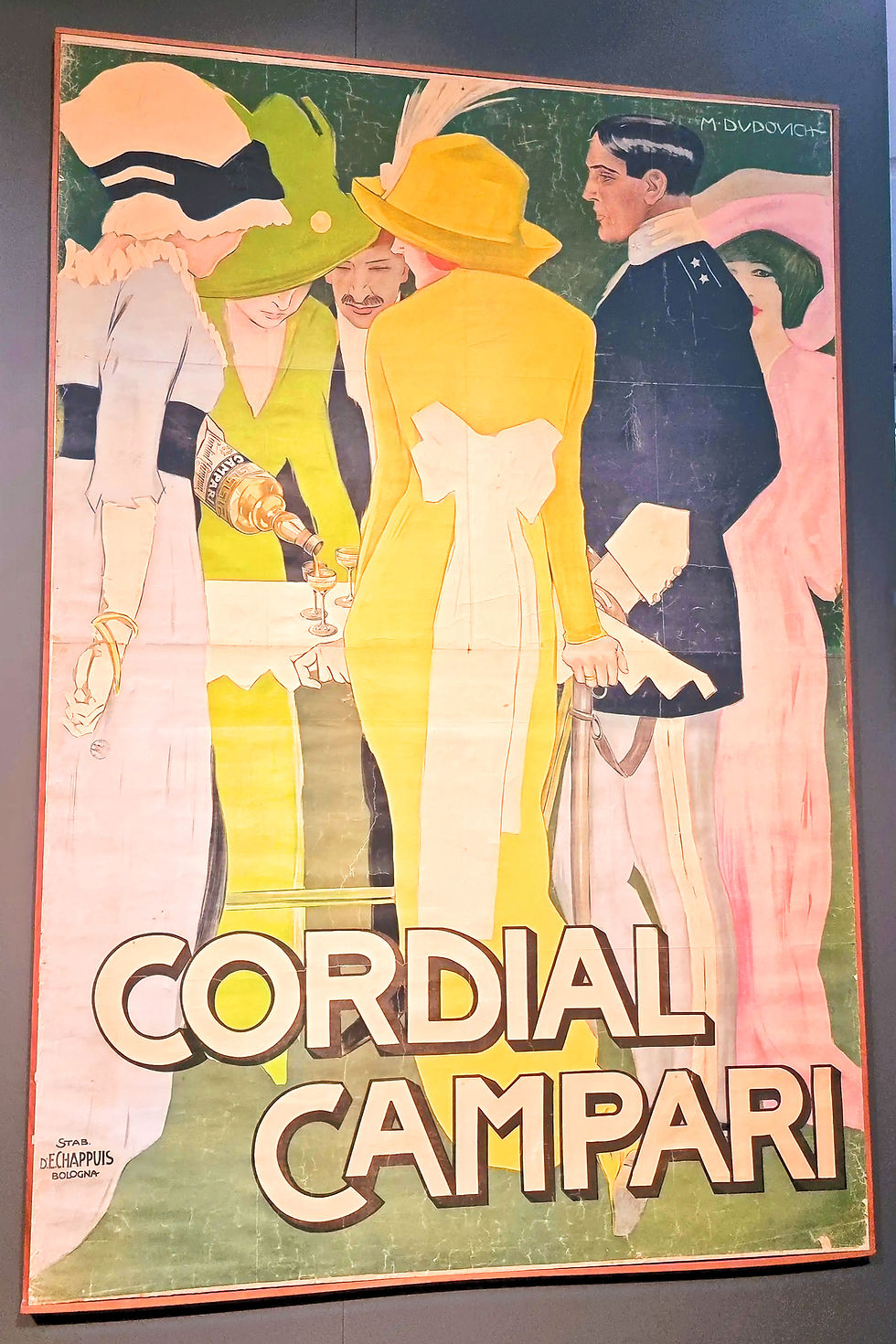 Five elegantly dressed people in hats gather around a table, with one pouring from a bottle labeled Campari. Text reads "Cordial Campari." Poster from Galleria Campari in Milan.