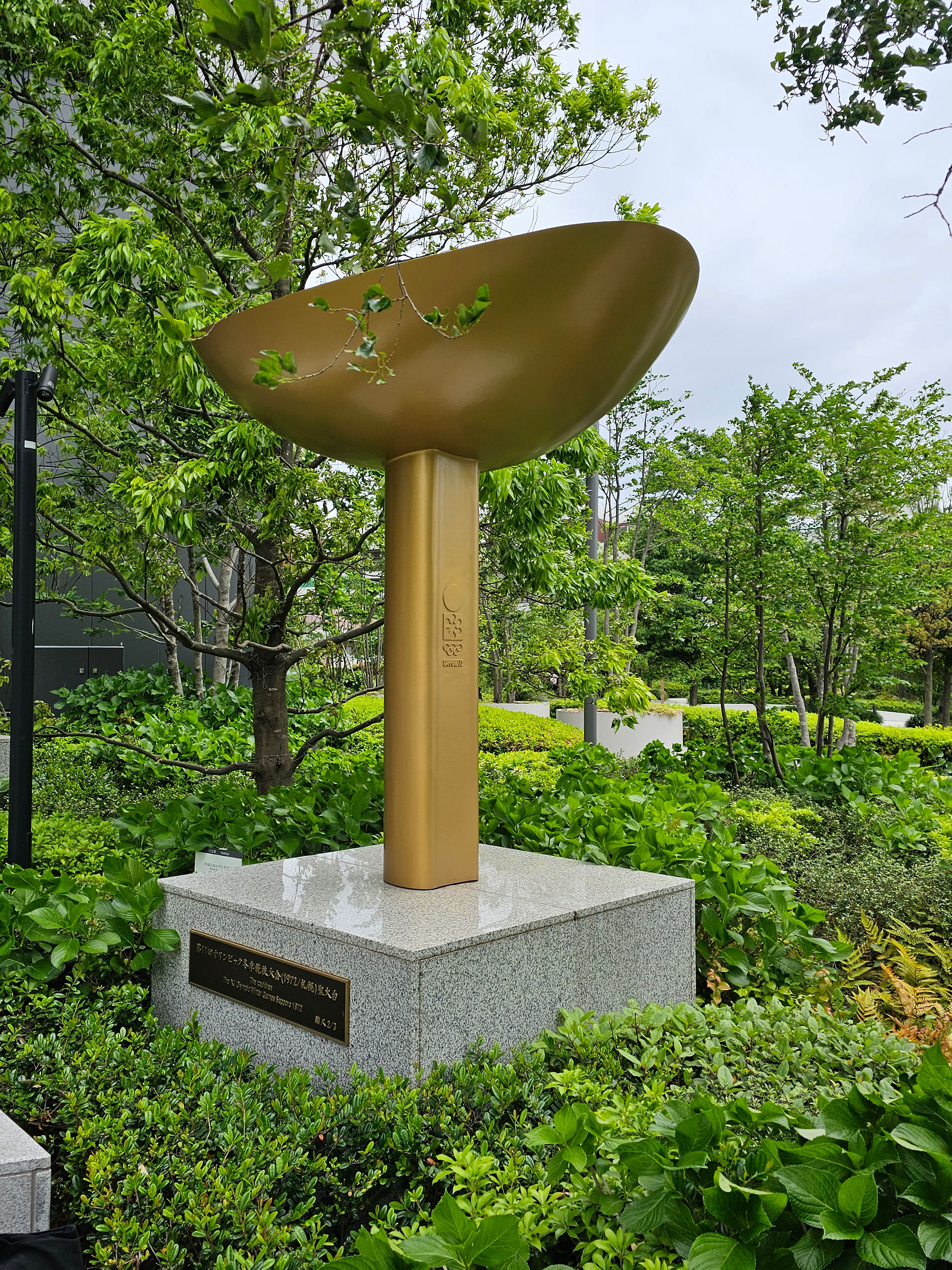 a gold olympic flame statue sits on a pale grey stone plinth surrounded by greenery. the gold statue has a thin vertical base and an open bowl top