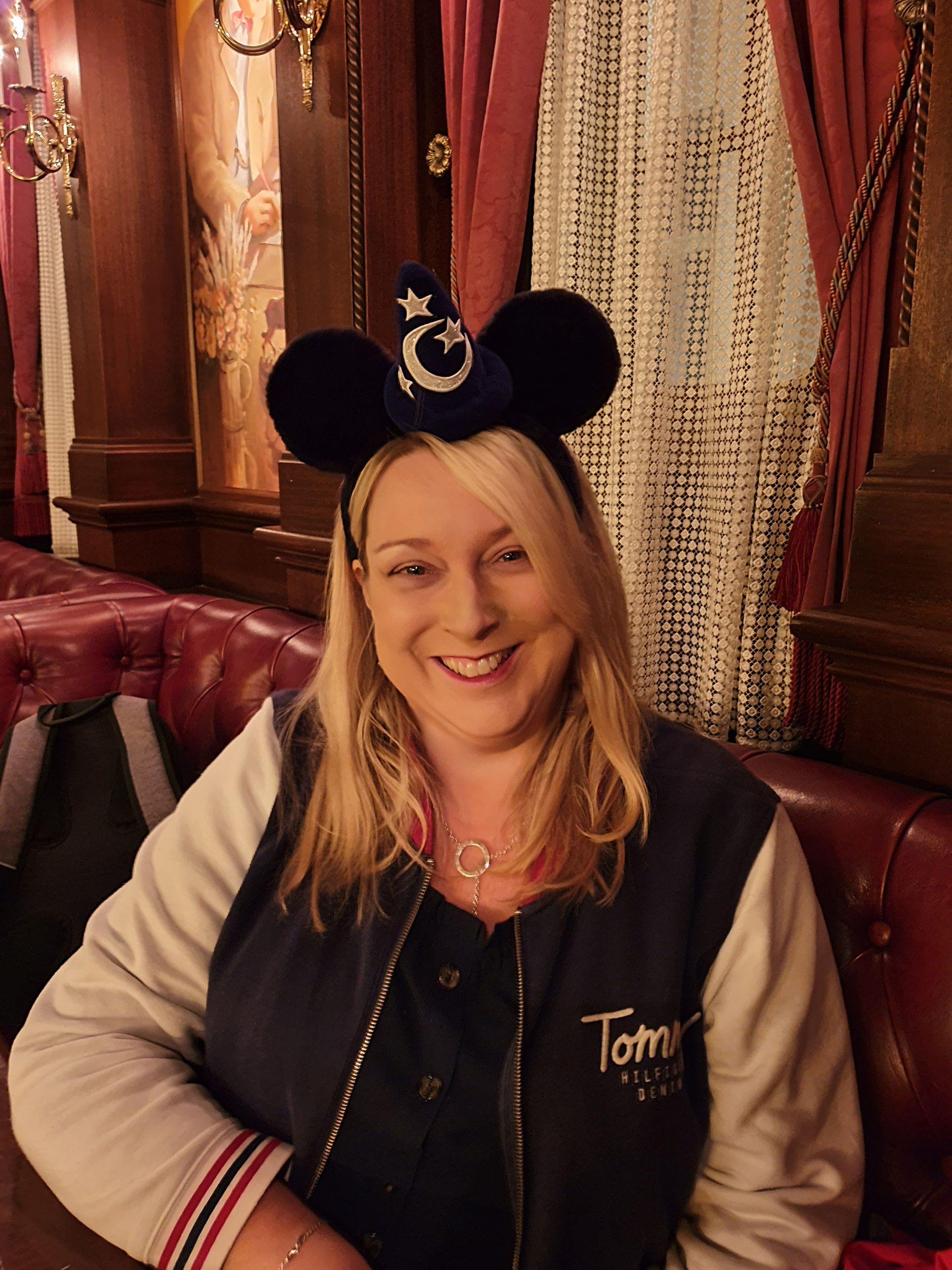 woman with blonde hair wears a bomber jacket and smiles as she wears mickey mouse ears headband sitting in a red leather button chair inside the SS Columbia steam liner in the American waterfront land at Tokyo Disneysea.
