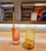 Two cocktails on a wooden bar, one pink with orange garnish and straw, the other yellow with signage for the brand at the johnnie walker princes street brand home. Shelves with glasses and bottles in the background.