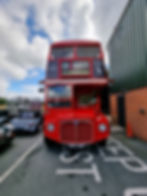 A classic red double-decker bus labeled "159 Sloane Square" is parked beside a building under a partly cloudy sky. Other cars are visible nearby.