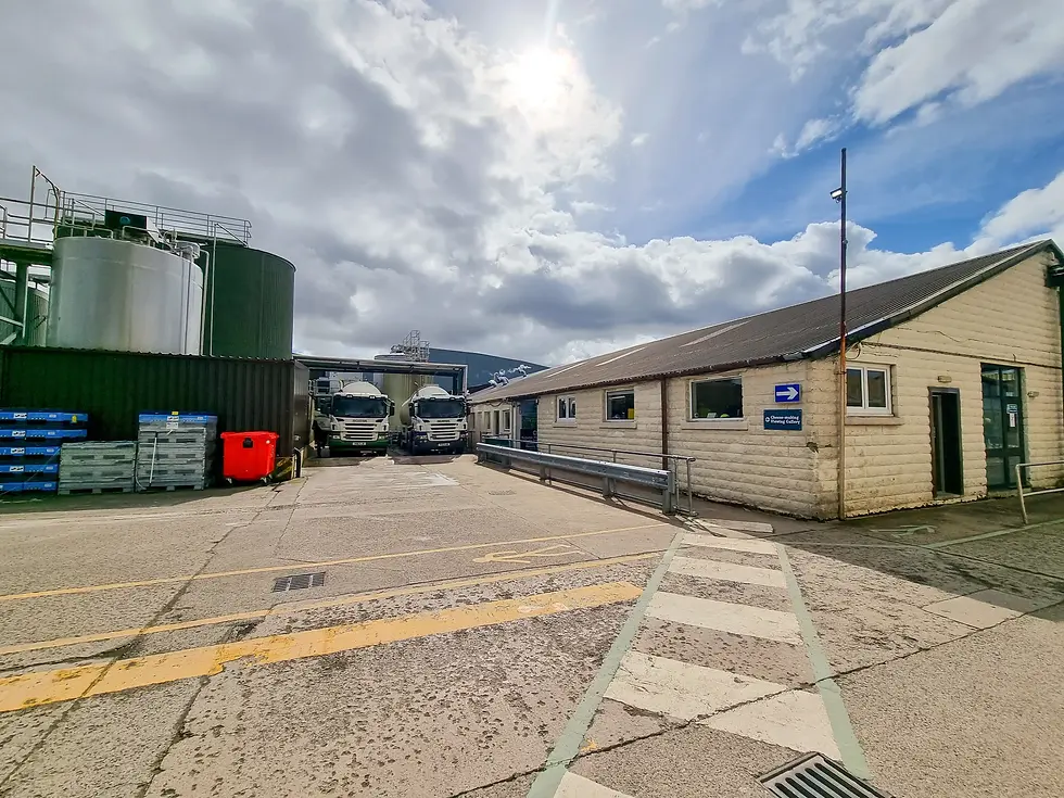 Industrial yard at wensleydale creamery with two trucks near metal tanks and a beige building. Blue sky with clouds, yellow lines, and a red bin visible.