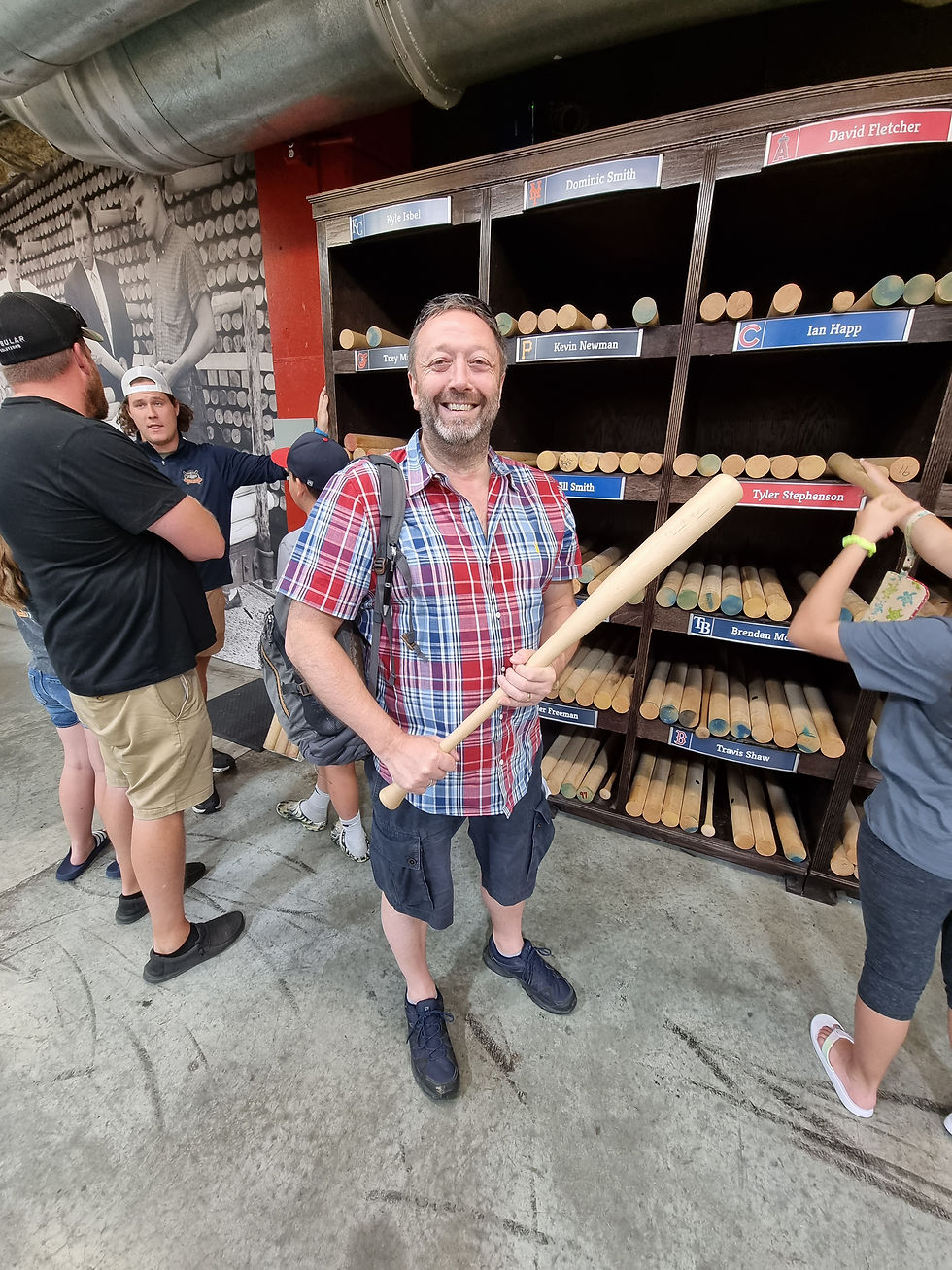 Man smiling, holding a baseball bat in front of a wooden rack with labeled bats. Casual setting, people interacting, checkered shirt.
