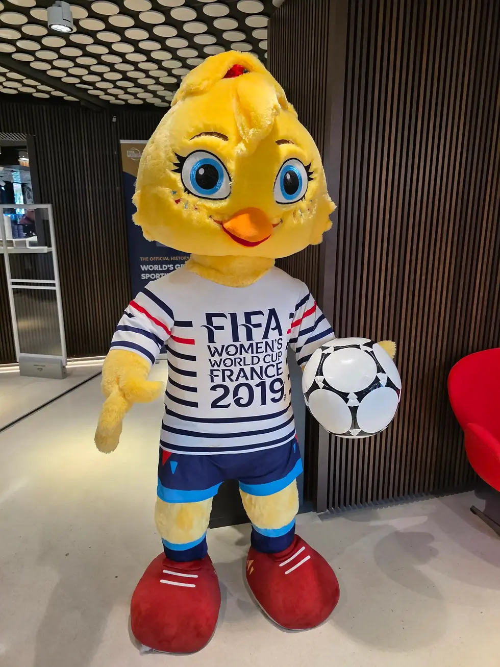 Yellow chick mascot in FIFA Women’s World Cup France 2019 shirt holds a soccer ball in an indoor display, smiling brightly.