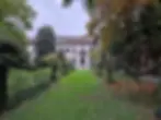 Pathway through a lush garden leads to a historic white villa with arched windows. Greenery surrounds, creating a tranquil, serene vibe.