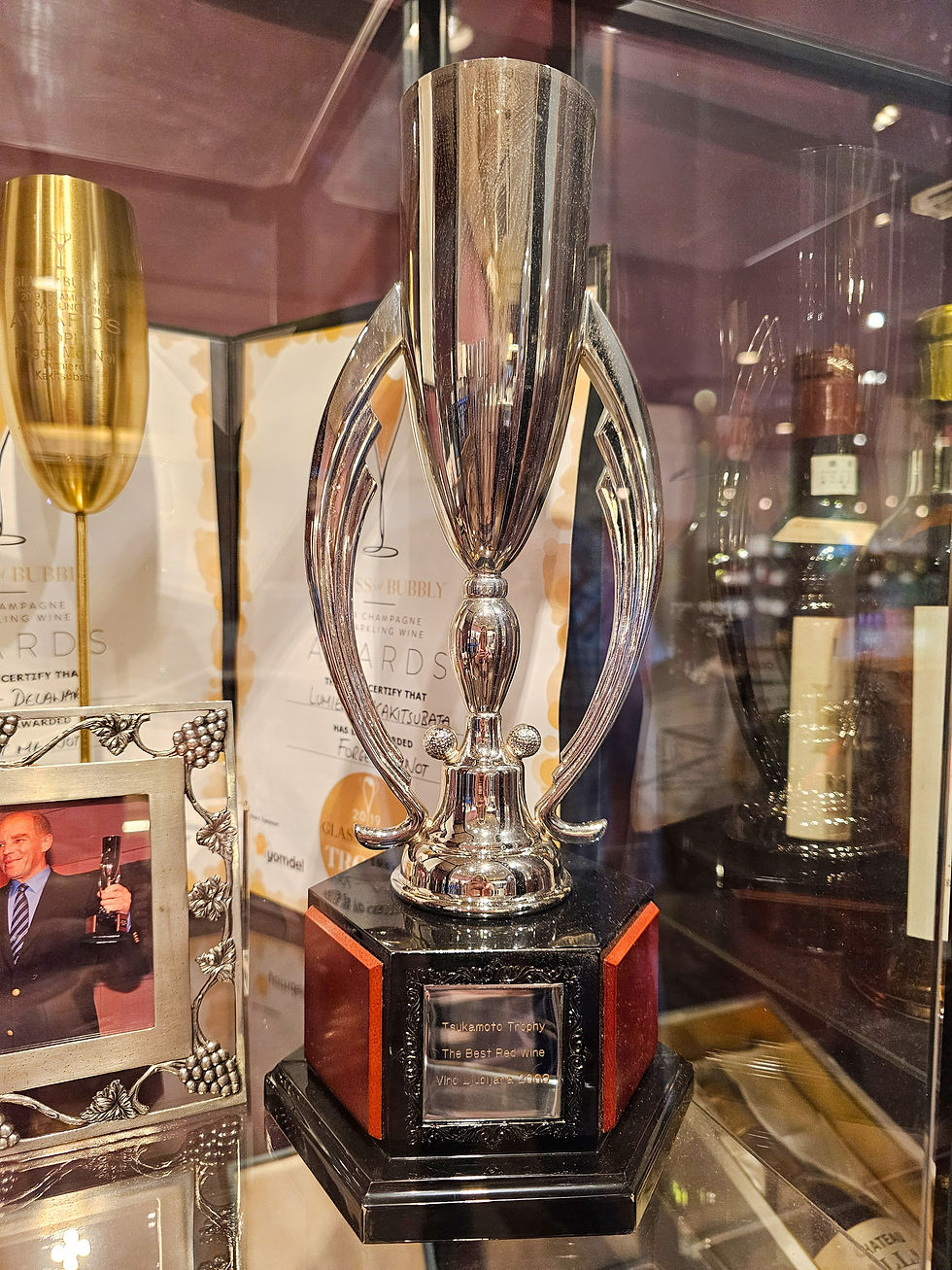 Silver trophy in a glass display case, flanked by gold awards and wine bottles; plaque reads Tsukamoto Trophy, The Best Red Wine.