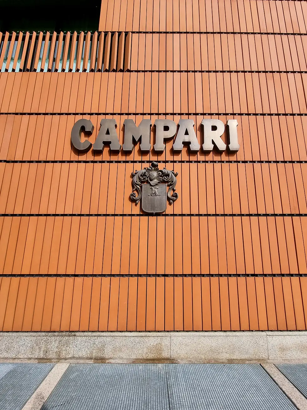 Orange brick building facade with bold "CAMPARI" text and ornate emblem. Vertical stripes add texture. Ground appears stone and metal. Exterior of the Galleria Campari in Milan.
