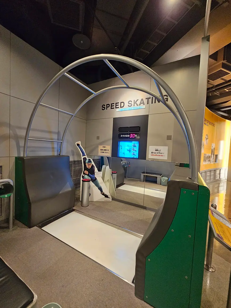 Speed skating simulation display at the Sapporo Olympic Museum in Japan, with life-size skater cutout, gray walls, and green accents. Text reads "Speed Skating" and "30."