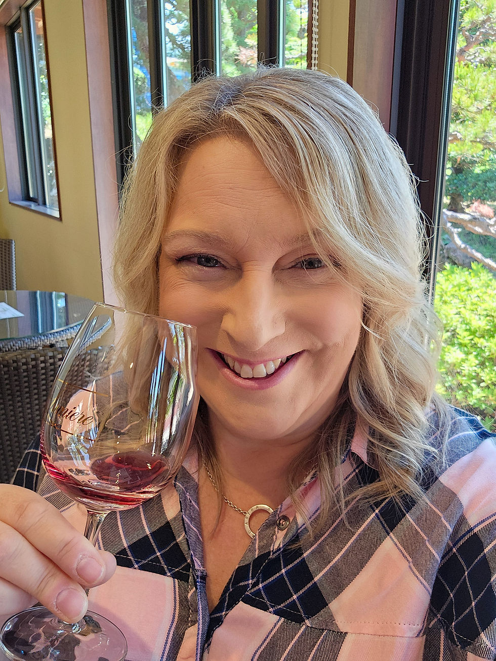 Smiling blonde woman in a plaid shirt holds a wine glass in a bright room with large windows and greenery outside.