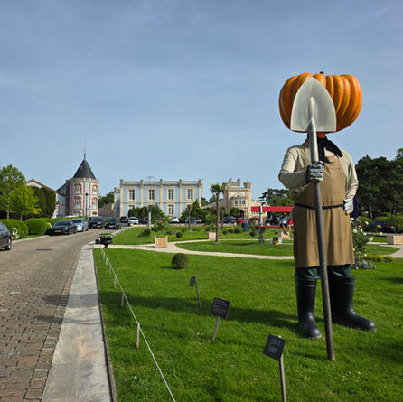 Giant pumpkin-headed scarecrow statue with shovel on a manicured lawn, beside chateau-like buildings and a Dont touch sign
