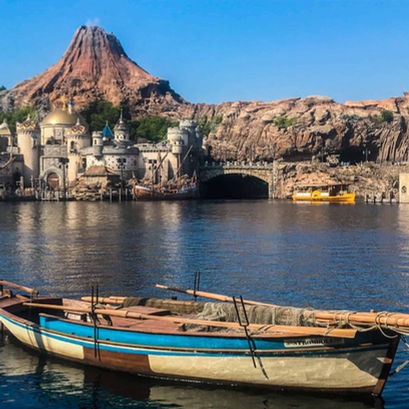 A wooden boat floats on calm water. In the background, a castle-like structure sits beneath a rugged mountain under a clear blue sky. This is the scene at Tokyo disneysea, the disney brand's japanese visitor experience