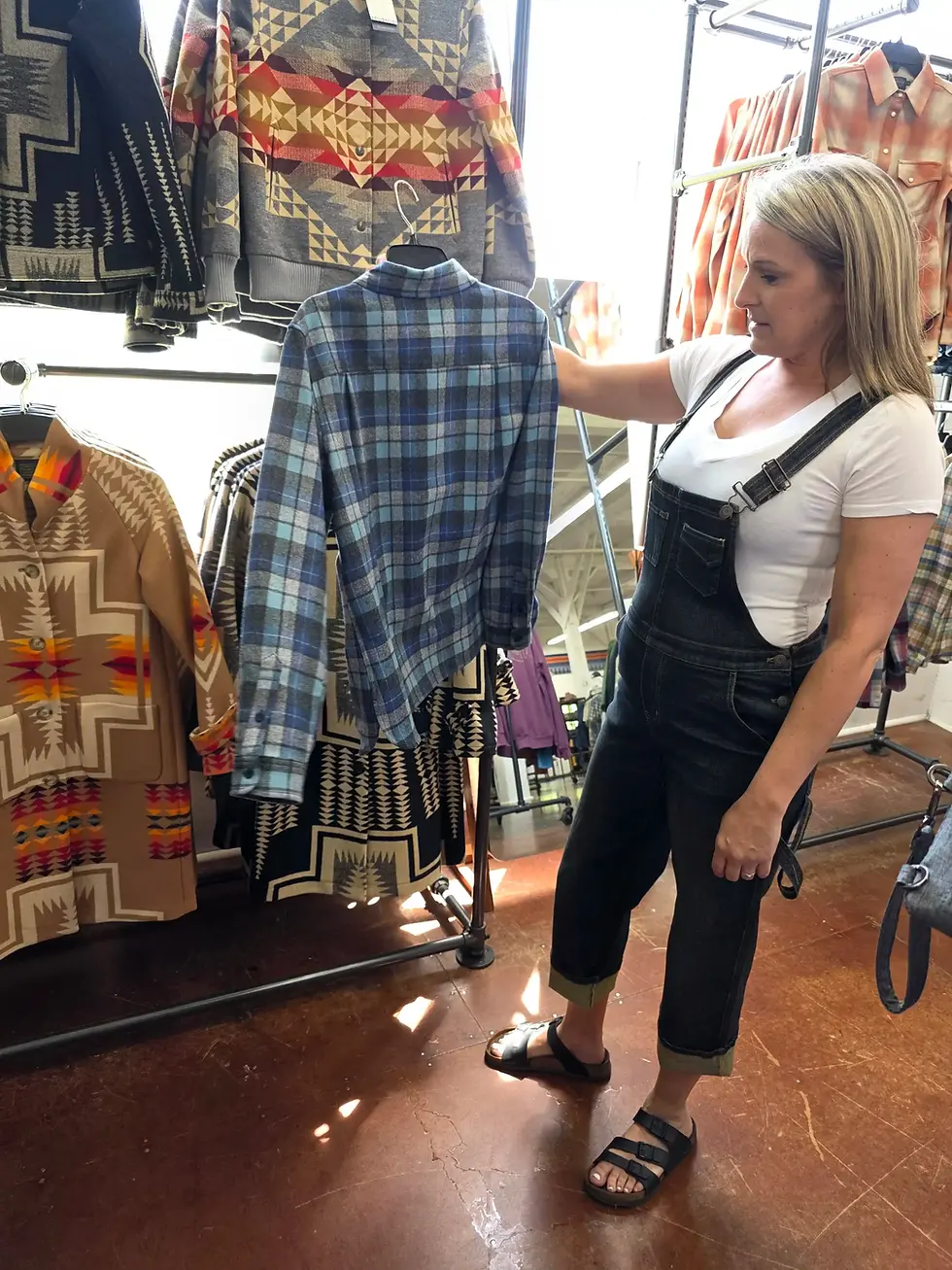 A woman in denim overalls holds a blue plaid shirt in a clothing store with patterned garments on racks nearby, sunlit floor at the shop for the Pendleton Woolen Mill Tour Washougal.