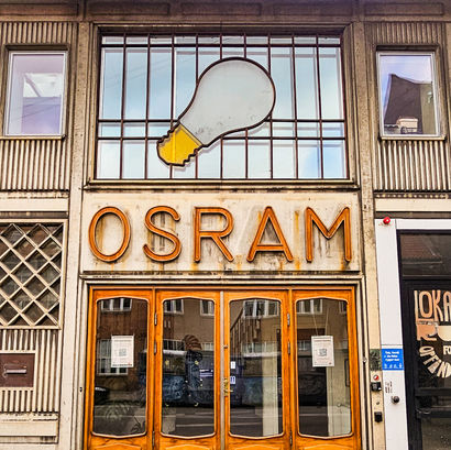 Art Deco building with "OSRAM" sign, large light bulb art above wooden doors. Reflections of buildings in glass, striped wall pattern.