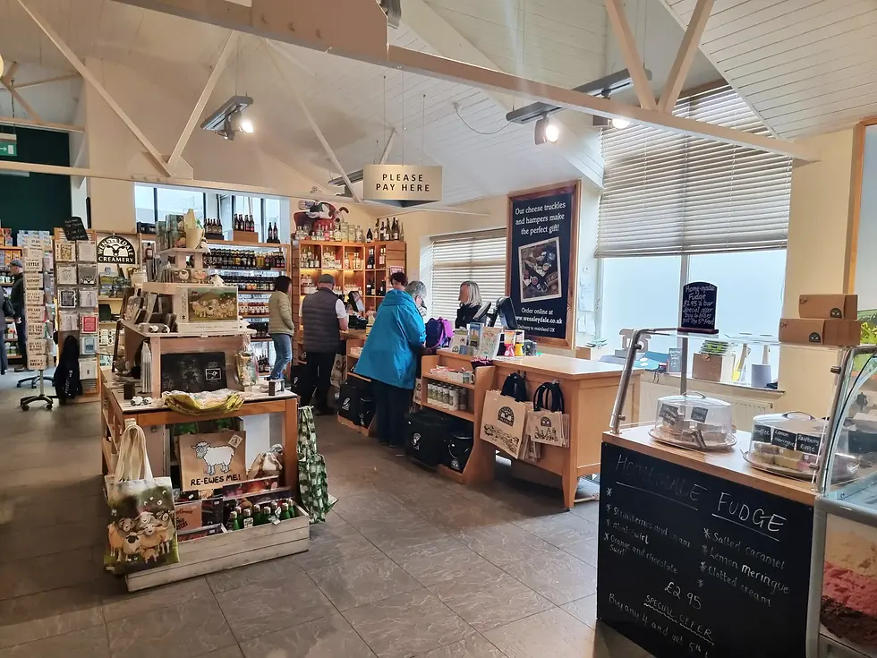 Shop interior at wensleydale creamery with people at a checkout, shelves filled with products. Sign reads "Please Pay Here." A board lists types of homemade fudge.