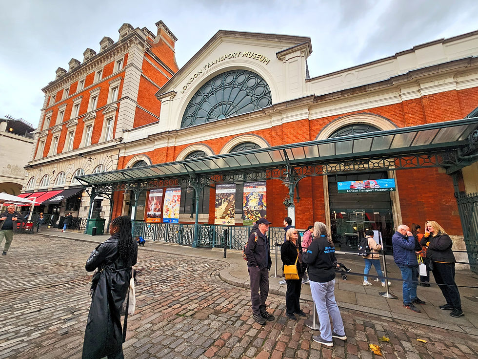 People gathered by London Transport Museum entrance, chatting. Historic red-brick building. Overcast sky. Museum signs visible.