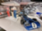 Blue race car with number 2 in the Silverstone museum. Nearby, vintage racing suits displayed in a glass case. Black-and-white race scene mural in background.