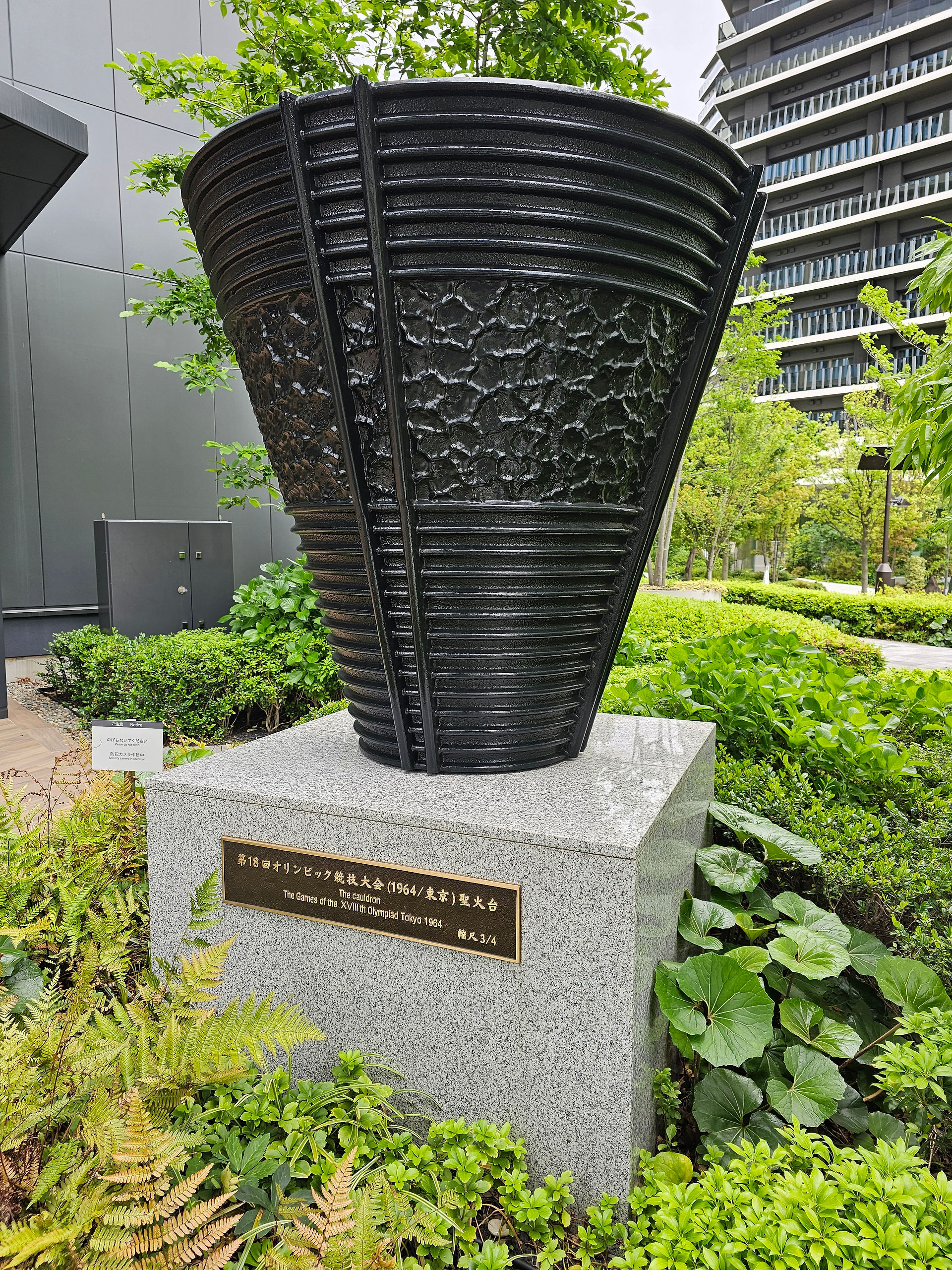 black metal upturned cone shaped olympic flame statue on a pale grey stone plinth surrounded by green plants