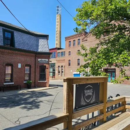 Brick brewery buildings under a clear blue sky, sign reads Samuel Adams. "Haffenreffer" on yellow smokestack. Tree, bench, and urban setting in Boston at the Samuel Adams Brewery tour.