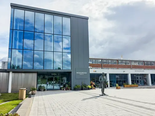 World of Wedgwood's modern building with large glass windows reflecting clouds. "Wedgwood" sign visible. Bronze statue stands in front on a paved walkway.