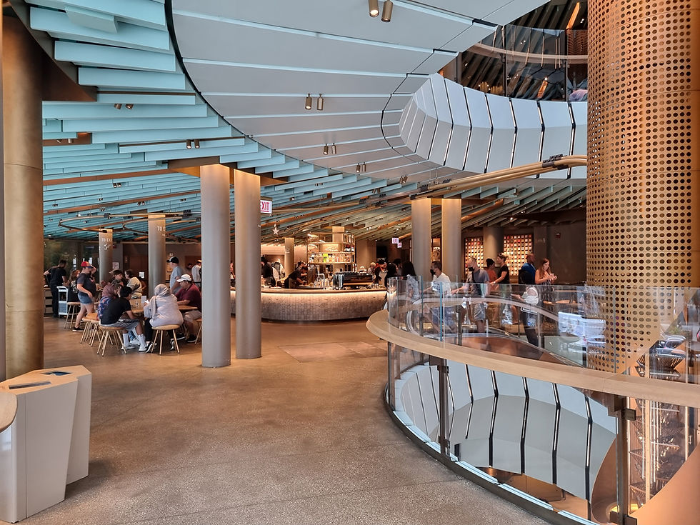 Modern cafe with people seated at tables, others queuing at a curved bar. Blue and gold decor, spiral staircase, and "Exit" sign visible.