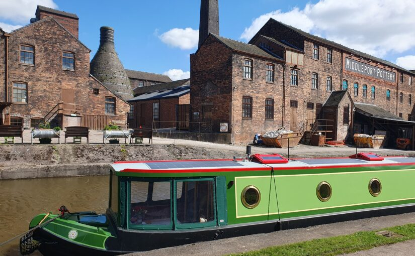 Green canal boat moored by Middleport Pottery, old brick buildings in the background. Sunny day, blue sky with few clouds.