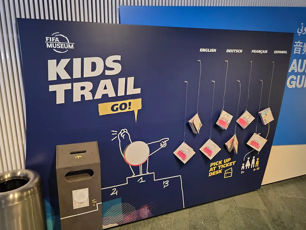 FIFA Museum Kids Trail display with hanging booklets, language options, and a pickup sign on a blue wall in a museum lobby.
