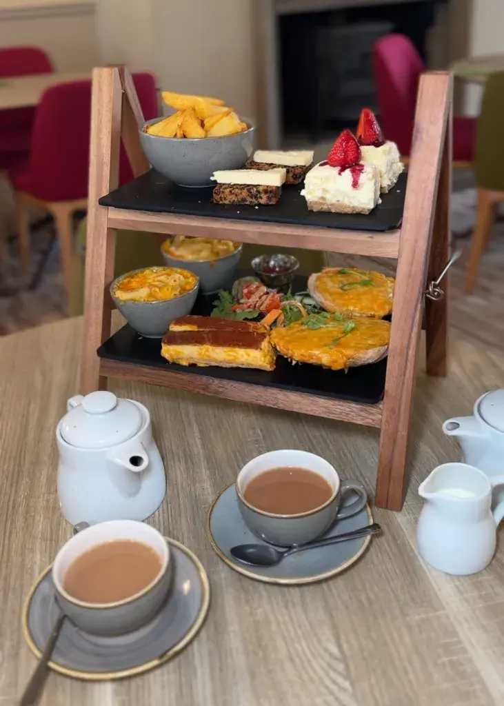 Scones, pastries, fries, and quiche are displayed on a wooden stand. Two cups of tea and a teapot sit on a table in a cozy cafe at wensleydale creamery.