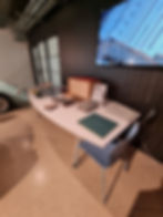 A table with car brochures and materials, wooden box, and a blue chair in an industrial space. Screen displays "Morgan Motor Company."