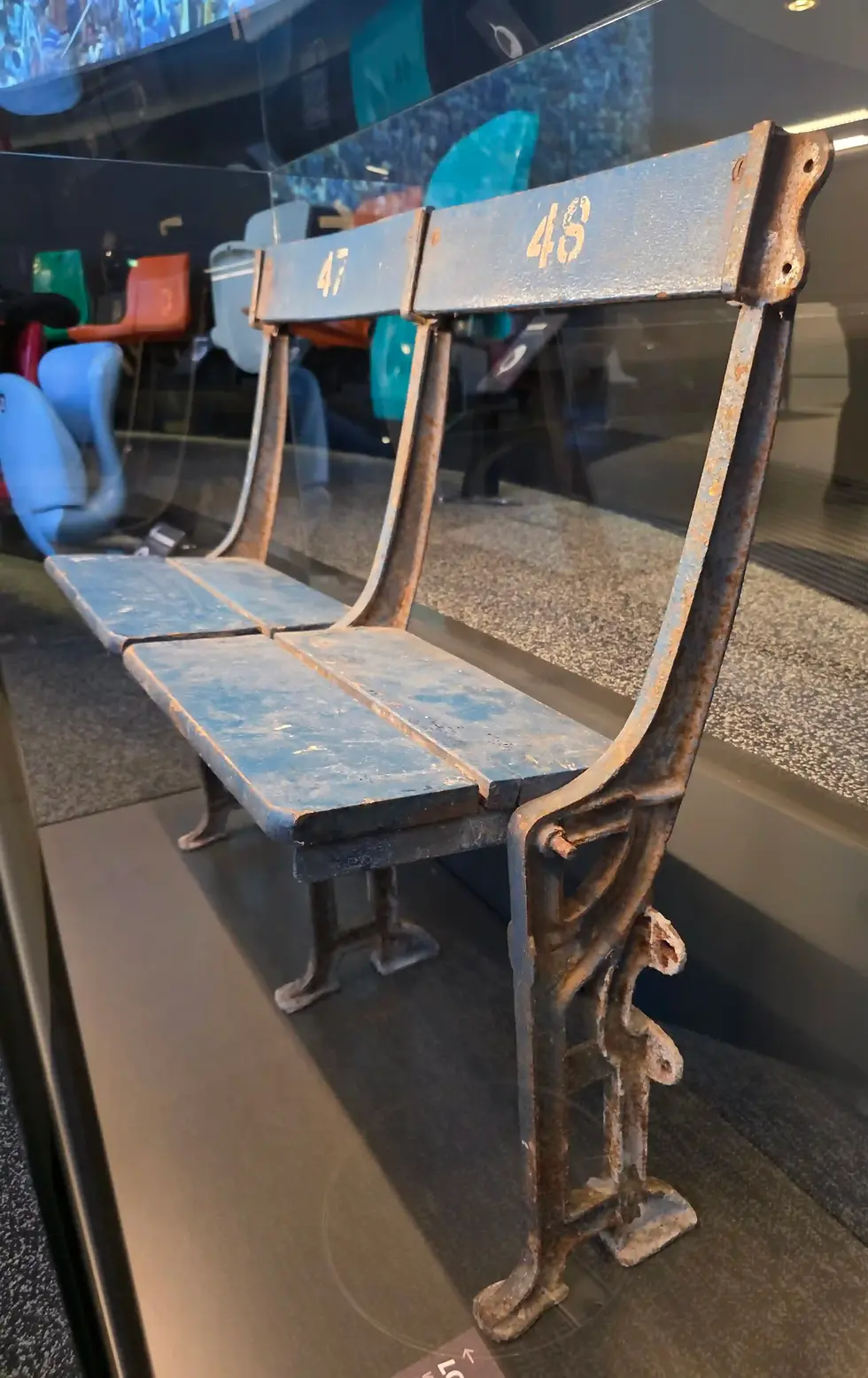 Rusty blue stadium bench seats numbered 47 and 48 in a museum display case, with reflections and colorful seats behind them at FIFA Museum.
