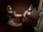 Copper distillery equipment in a dimly lit rustic stone room. Two copper stills and wooden barrels create an antique, industrial atmosphere.