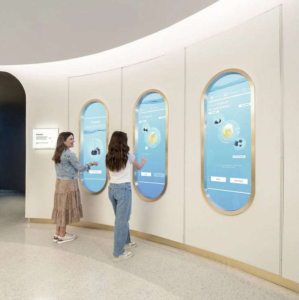 Two people interact with touchscreen panels displaying "Create your own flavor." The setting at World of Coca-Cola is modern and bright with large screens.